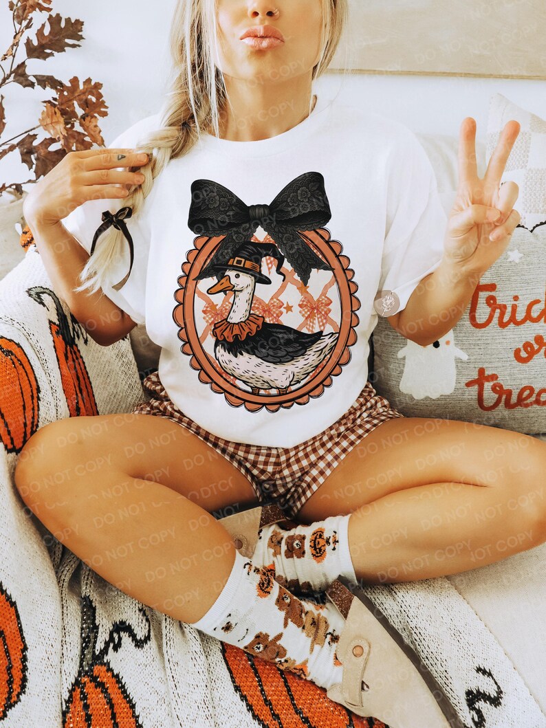 Peut inclure: T-shirt blanc avec un dessin d'oie de dessin anim&eacute; portant un chapeau de sorci&egrave;re et un n&oelig;ud noir. L'oie est dans un cadre ovale avec un motif de n&oelig;uds et d'&eacute;toiles. Le mod&egrave;le porte un short &agrave; carreaux marron et blanc et des chaussettes avec des graphiques d'ours.