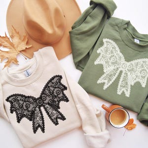 May include: Two sweatshirts, one cream and one olive green, each featuring a large lace bow design. A tan hat and a fall leaf are also in the image. A small brown mug with a drink is also present.