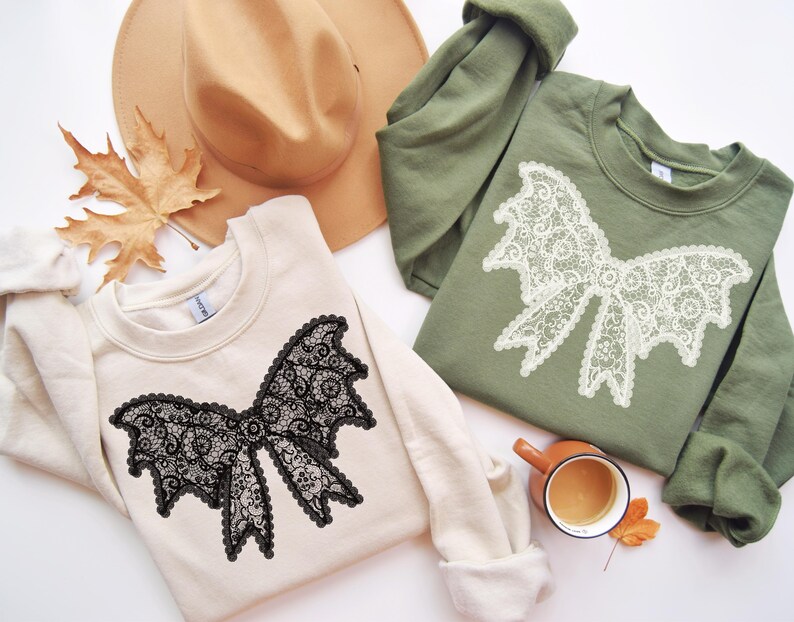 May include: Two sweatshirts, one cream and one olive green, each featuring a large lace bow design. A tan hat and a fall leaf are also in the image. A small brown mug with a drink is also present.