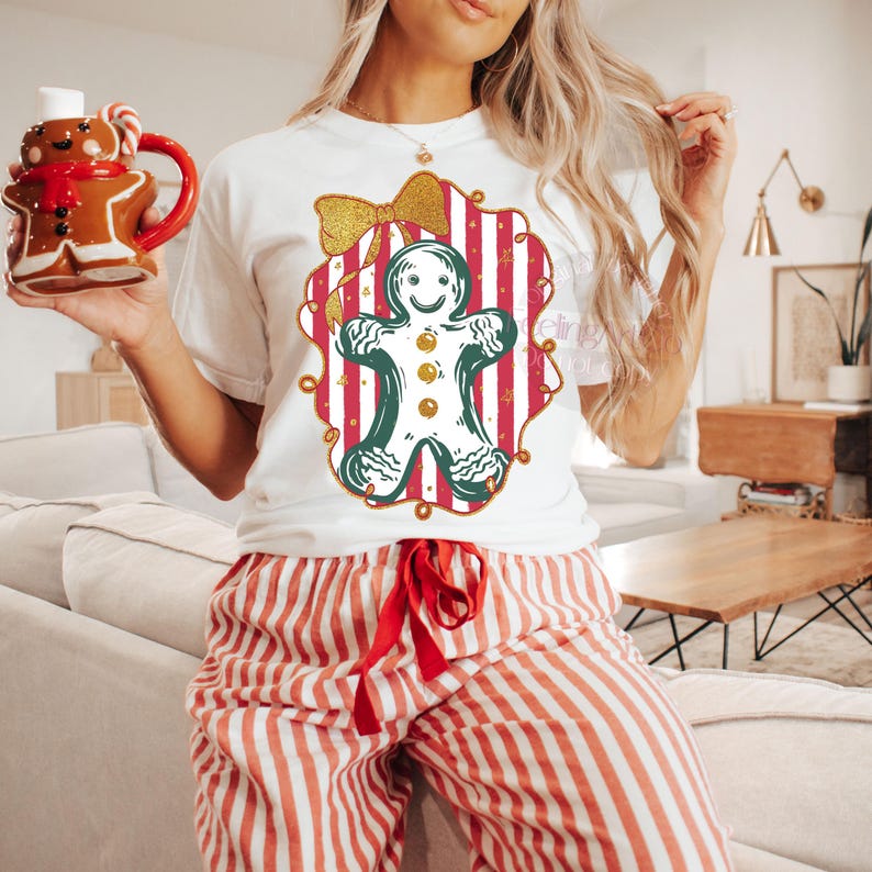 May include: A white t-shirt featuring a gingerbread man design with a gold bow and red and white striped background. The person is wearing red and white striped pajama pants and holding a gingerbread man mug.