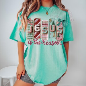 May include: A turquoise short-sleeved t-shirt with the words "JESUS is the reason" in a decorative font. The word "JESUS" is made up of colorful, stitched blocks with Christmas-themed designs.
