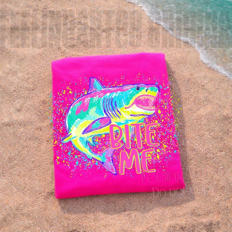 Op de afbeelding: Een opgevouwen felroze t-shirt met een kleurrijke haai-afbeelding. De haai is in tinten blauw, groen en geel, met de woorden "Bite Me" in roze en geel. Het shirt ligt op een zandstrand.