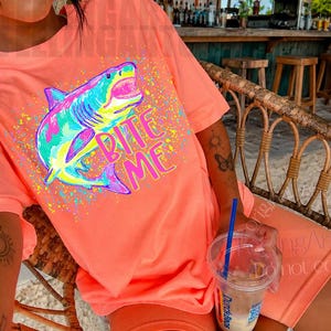 Op de afbeelding: Koraalrood t-shirt en short set met een kleurrijke haai afbeelding en de tekst "Bite Me". De haai is afgebeeld in blauw, groen, roze en geel, met kleurrijke verfspatten rond de afbeelding.