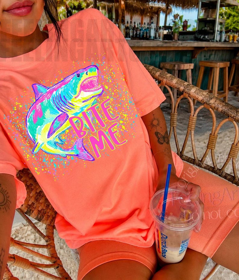 Op de afbeelding: Koraalrood t-shirt en short set met een kleurrijke haai afbeelding en de tekst "Bite Me". De haai is afgebeeld in blauw, groen, roze en geel, met kleurrijke verfspatten rond de afbeelding.