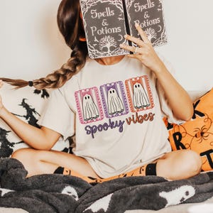 May include: A beige t-shirt with a Halloween-themed design. The shirt features three ghost illustrations with bows, framed in decorative borders, and the words "Spooky Vibes" in a purple script. The person is holding a book with the words "Spells & Potions".