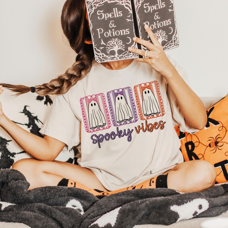 May include: A beige t-shirt with a Halloween-themed design. The shirt features three ghost illustrations with bows, framed in decorative borders, and the words "Spooky Vibes" in a purple script. The person is holding a book with the words "Spells & Potions".