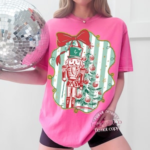 May include: Pink t-shirt with a Christmas design. The graphic shows a red nutcracker, a Christmas tree with baubles, and a red bow. The text "FeelingArtCo Do not copy" is printed on the shirt.