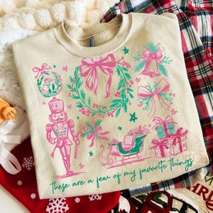 May include: Cream-colored sweatshirt with a festive design. The graphic includes a nutcracker, bows, a wreath, a sleigh, and gifts in pink and teal. The text "These are a few of my favorite things" is printed at the bottom.