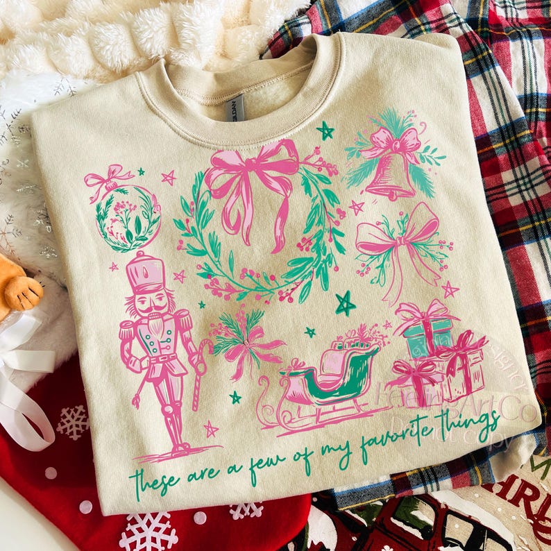 May include: Cream-colored sweatshirt with a festive design. The graphic includes a nutcracker, bows, a wreath, a sleigh, and gifts in pink and teal. The text "These are a few of my favorite things" is printed at the bottom.