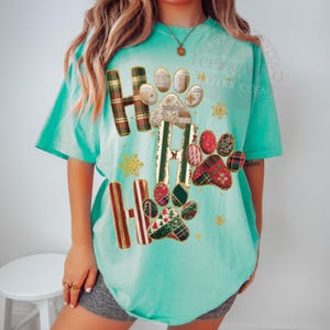 K&ouml;nnte beinhalten: T&uuml;rkisfarbenes T-Shirt mit festlichem Design. Das Shirt zeigt die Worte "HO HO HO" mit Pfotenabdr&uuml;cken und Schneeflocken. Die Buchstaben sind in einem Karomuster gehalten, und die Pfotenabdr&uuml;cke haben ein Patchwork-Design mit weihnachtlichen Mustern.