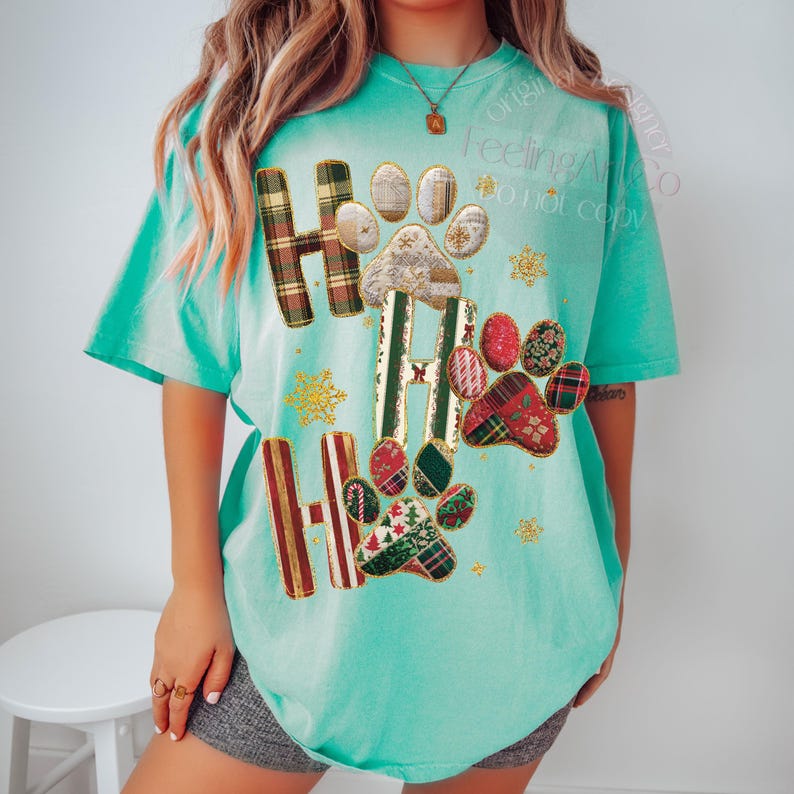 K&ouml;nnte beinhalten: T&uuml;rkisfarbenes T-Shirt mit festlichem Design. Das Shirt zeigt die Worte "HO HO HO" mit Pfotenabdr&uuml;cken und Schneeflocken. Die Buchstaben sind in einem Karomuster gehalten, und die Pfotenabdr&uuml;cke haben ein Patchwork-Design mit weihnachtlichen Mustern.