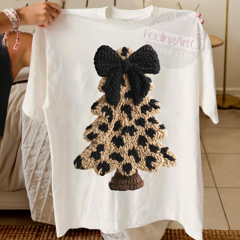 May include: White t-shirt featuring a knitted Christmas tree design. The tree has a tan base with black spots and a black bow at the top. The tree trunk is brown. The shirt is held up by a person.
