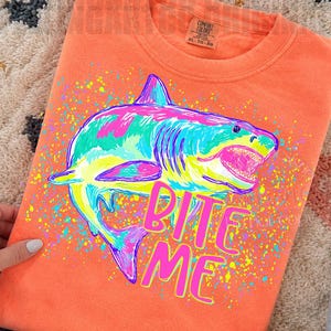 Op de afbeelding: Koraalrood sweatshirt met een kleurrijke haai-afbeelding. De haai is omlijnd in blauw, roze, geel en groen, met de tekst "BITE ME" in roze en geel. Het label is Comfort Colors XL.
