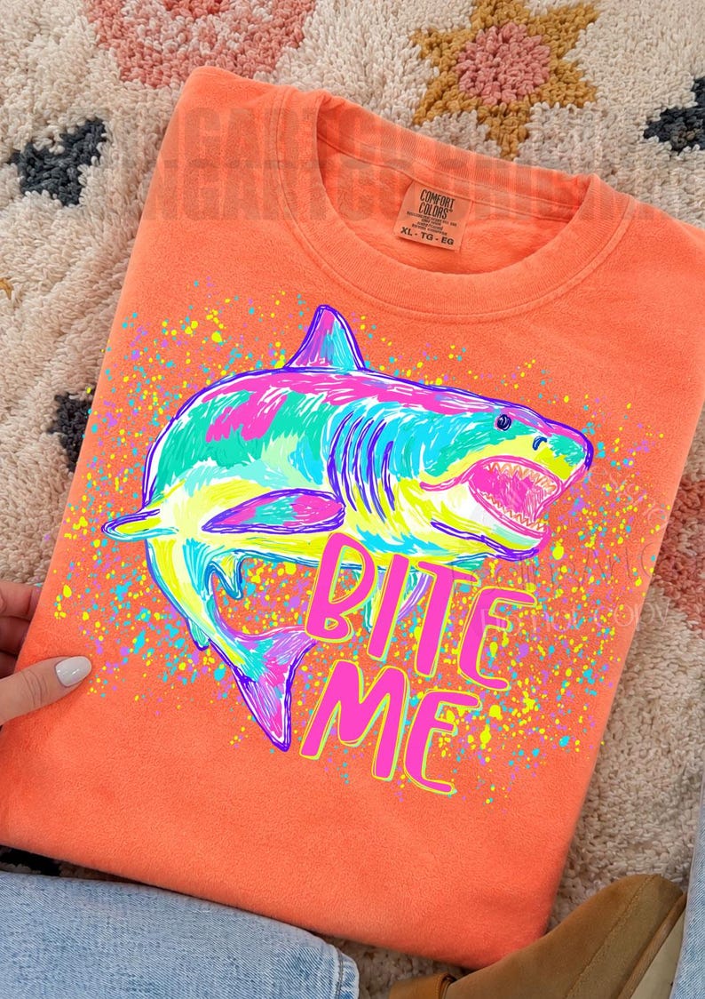 Op de afbeelding: Koraalrood sweatshirt met een kleurrijke haai-afbeelding. De haai is omlijnd in blauw, roze, geel en groen, met de tekst "BITE ME" in roze en geel. Het label is Comfort Colors XL.