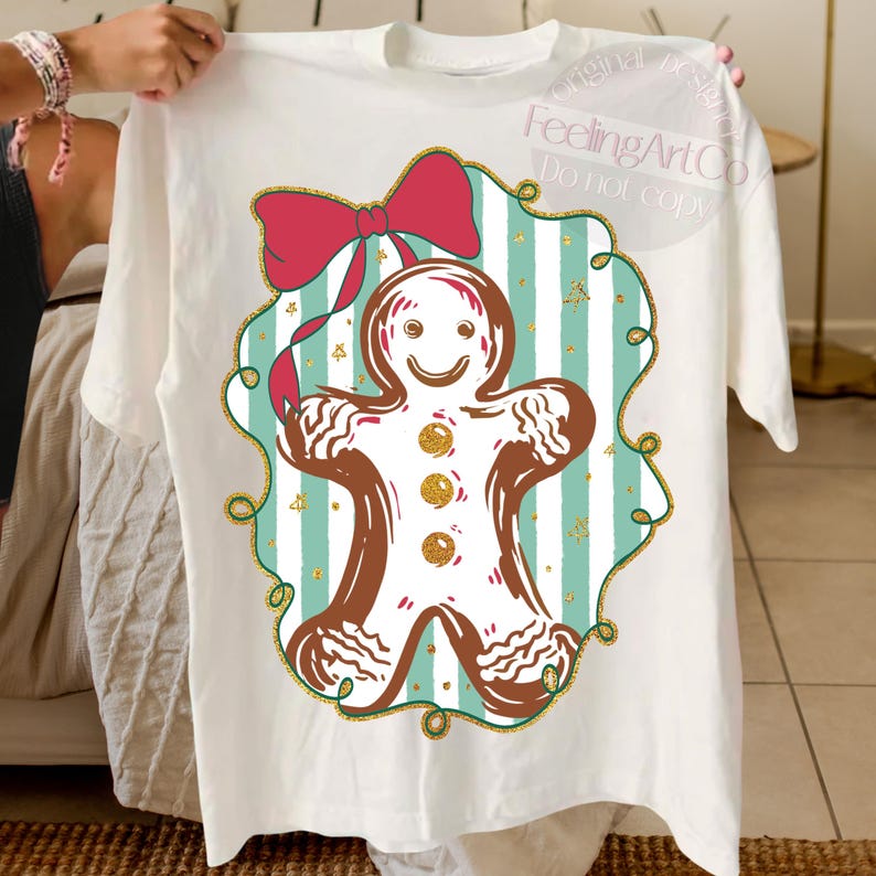 May include: White t-shirt featuring a gingerbread man design. The gingerbread man is brown with white icing details and gold accents. A red bow is at the top of the design, set against a teal and white striped background. The text "FeelingArtCo Do not copy" is visible.
