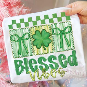 Op de afbeelding: Wit T-shirt met een St. Patrick's Day ontwerp. Het ontwerp bevat groene strikken, een klavertjevier en de woorden "Blessed Vibes" in groen en geel. Bovenop staat een groen-wit geblokt patroon.