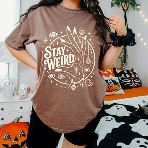 May include: Brown t-shirt with the text "Stay Weird" in a circular design featuring a hand, eyes, and celestial elements. The shirt is paired with black shorts. Halloween-themed pillows and decor are in the background.
