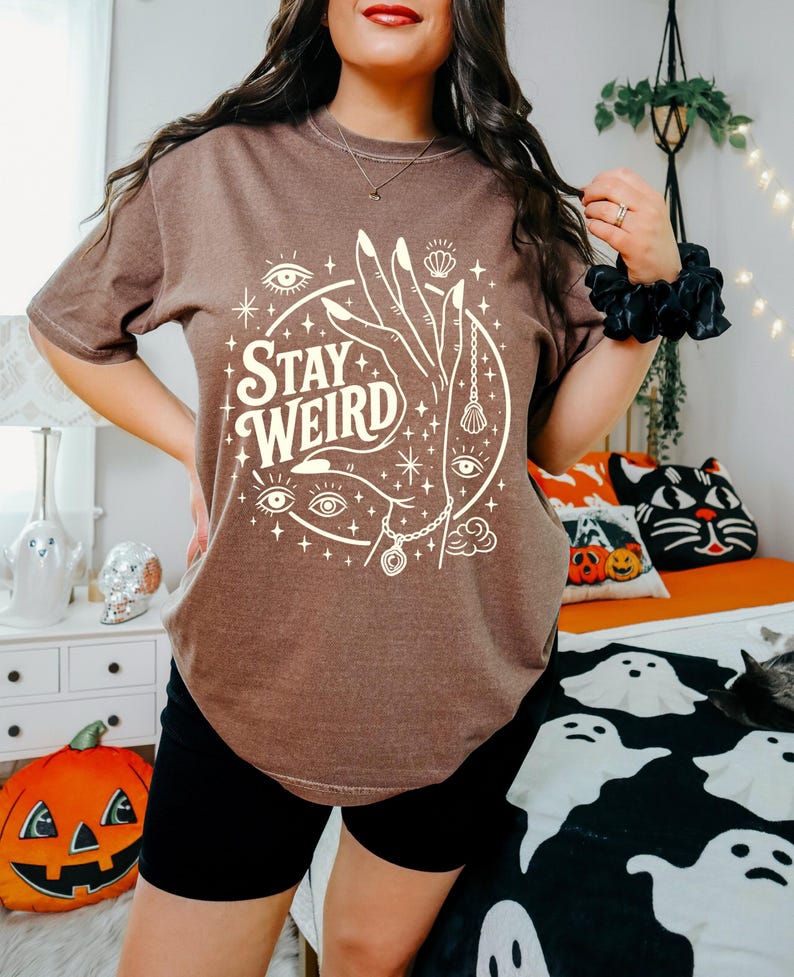 May include: Brown t-shirt with the text "Stay Weird" in a circular design featuring a hand, eyes, and celestial elements. The shirt is paired with black shorts. Halloween-themed pillows and decor are in the background.