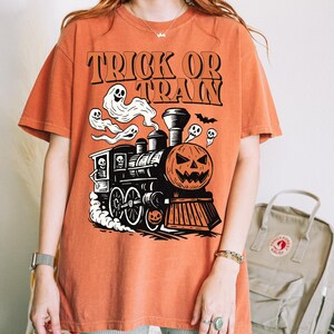 May include: Orange t-shirt with a Halloween train graphic. The design includes a pumpkin-faced train, ghosts, and the words "TRICK OR TRAIN" in black. The shirt is short-sleeved and casual.