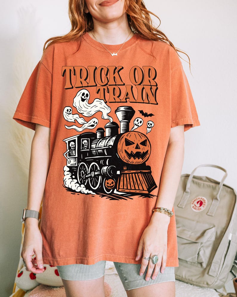 May include: Orange t-shirt with a Halloween train graphic. The design includes a pumpkin-faced train, ghosts, and the words "TRICK OR TRAIN" in black. The shirt is short-sleeved and casual.