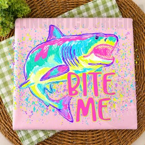 Op de afbeelding: Roze t-shirt met een kleurrijke haai-afbeelding. De haai is afgebeeld in tinten blauw, groen, roze en geel, met zijn bek open. De woorden "BITE ME" staan in felroze eronder.