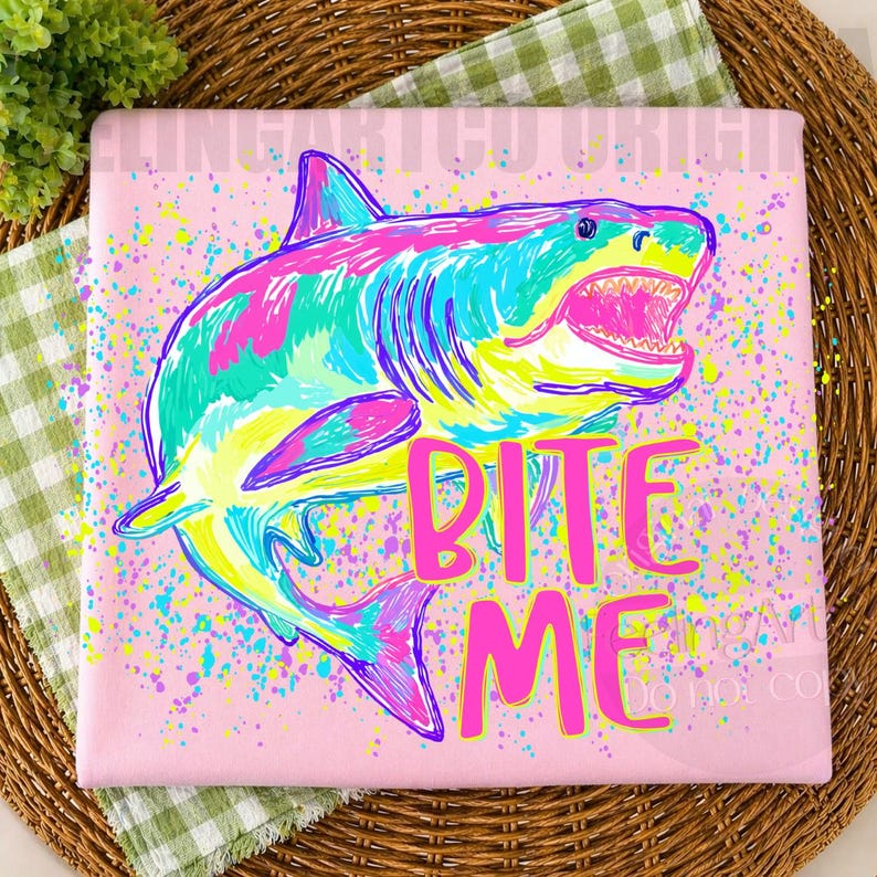 Op de afbeelding: Roze t-shirt met een kleurrijke haai-afbeelding. De haai is afgebeeld in tinten blauw, groen, roze en geel, met zijn bek open. De woorden "BITE ME" staan in felroze eronder.