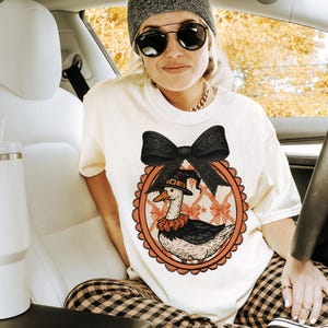 Peut inclure: T-shirt cr&egrave;me avec un dessin d'oie de dessin anim&eacute; portant un chapeau de sorci&egrave;re et un col &agrave; volants, dans un cadre ovale orange. Le motif comprend un grand n&oelig;ud noir. La personne porte un bonnet gris, des lunettes de soleil et une cha&icirc;ne dor&eacute;e.