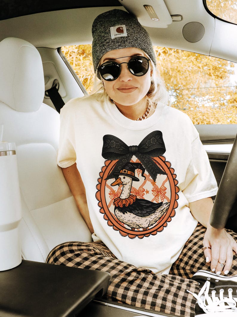 Peut inclure: T-shirt cr&egrave;me avec un dessin d'oie de dessin anim&eacute; portant un chapeau de sorci&egrave;re et un col &agrave; volants, dans un cadre ovale orange. Le motif comprend un grand n&oelig;ud noir. La personne porte un bonnet gris, des lunettes de soleil et une cha&icirc;ne dor&eacute;e.