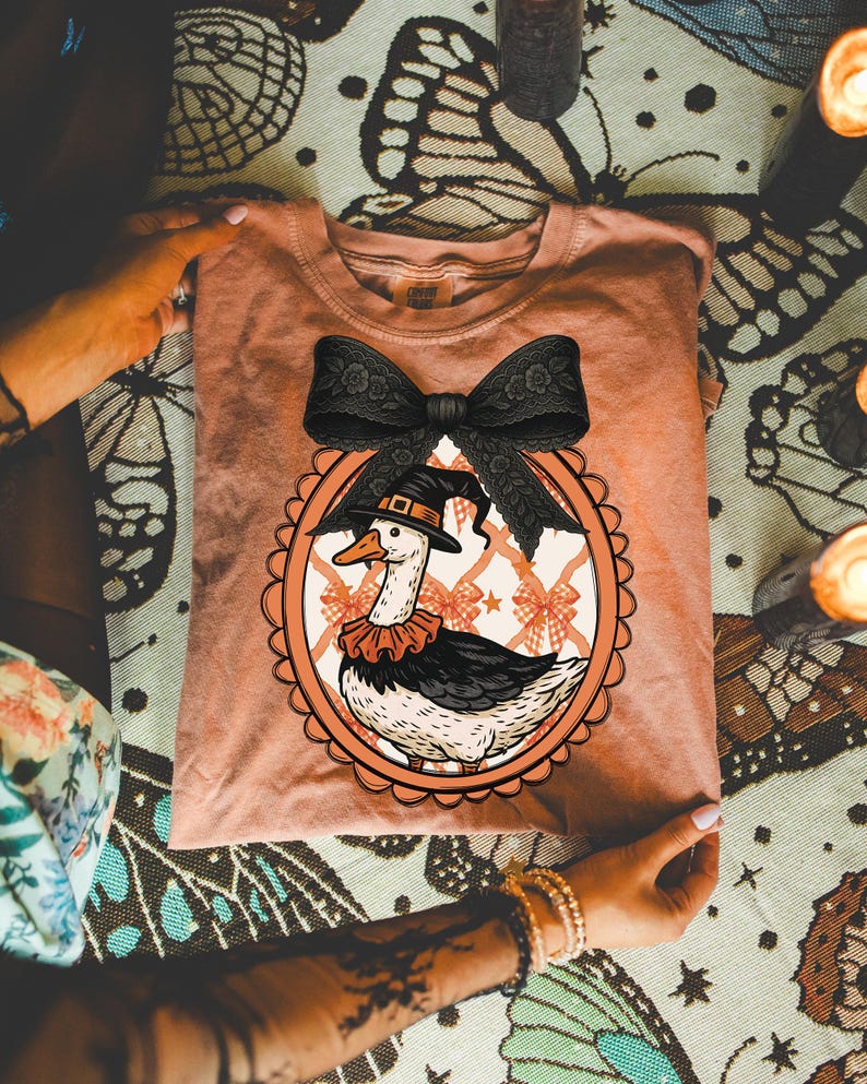 Peut inclure: Un t-shirt p&ecirc;che avec une illustration fantaisiste d'une oie portant un chapeau de sorci&egrave;re et un col &agrave; volants, encadr&eacute;e d'un n&oelig;ud noir. Le motif est ins&eacute;r&eacute; dans un cadre ovale avec un fond &agrave; motifs de n&oelig;uds. Le t-shirt est pli&eacute;.
