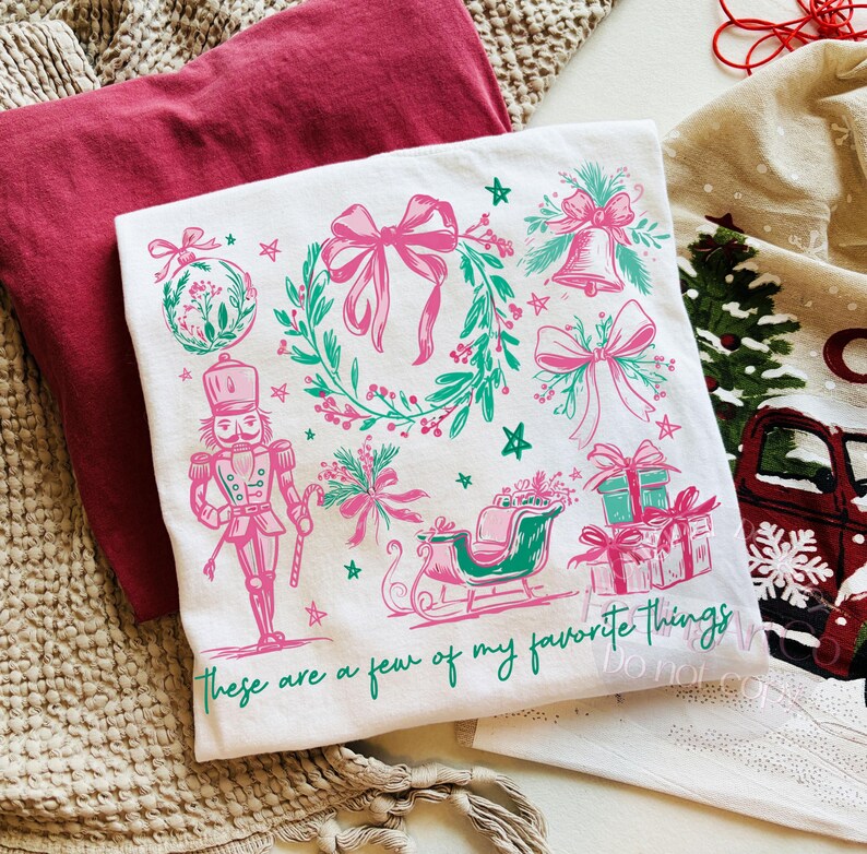 May include: White t-shirt with a pink and green Christmas design. The design includes a wreath, bows, a bell, a nutcracker, a sleigh, and gifts. The text "These are a few of my favorite things" is printed at the bottom.