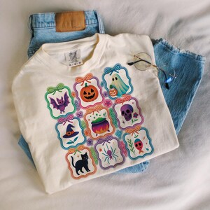 May include: Cream-colored t-shirt featuring a Halloween-themed design. The design includes various spooky illustrations such as a pumpkin, ghost, bat, skull, witch's hat, spider, and black cat. The shirt is paired with light blue denim jeans and round-framed glasses.