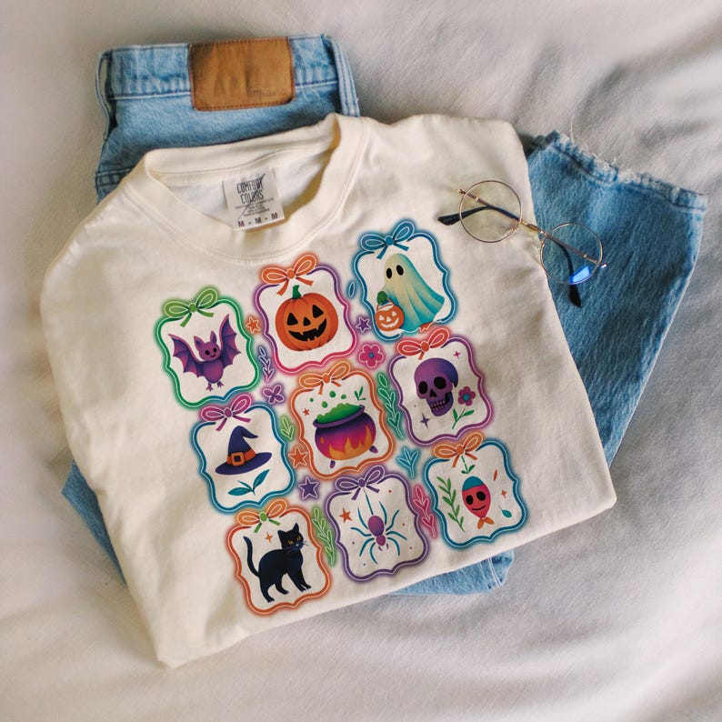 May include: Cream-colored t-shirt featuring a Halloween-themed design. The design includes various spooky illustrations such as a pumpkin, ghost, bat, skull, witch's hat, spider, and black cat. The shirt is paired with light blue denim jeans and round-framed glasses.