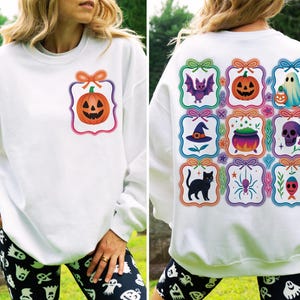 May include: White Halloween-themed sweatshirt with a pumpkin graphic on the front and various Halloween icons on the back. The back features a bat, pumpkin, ghost, witch hat, cauldron, skull, black cat, spider, and fish.