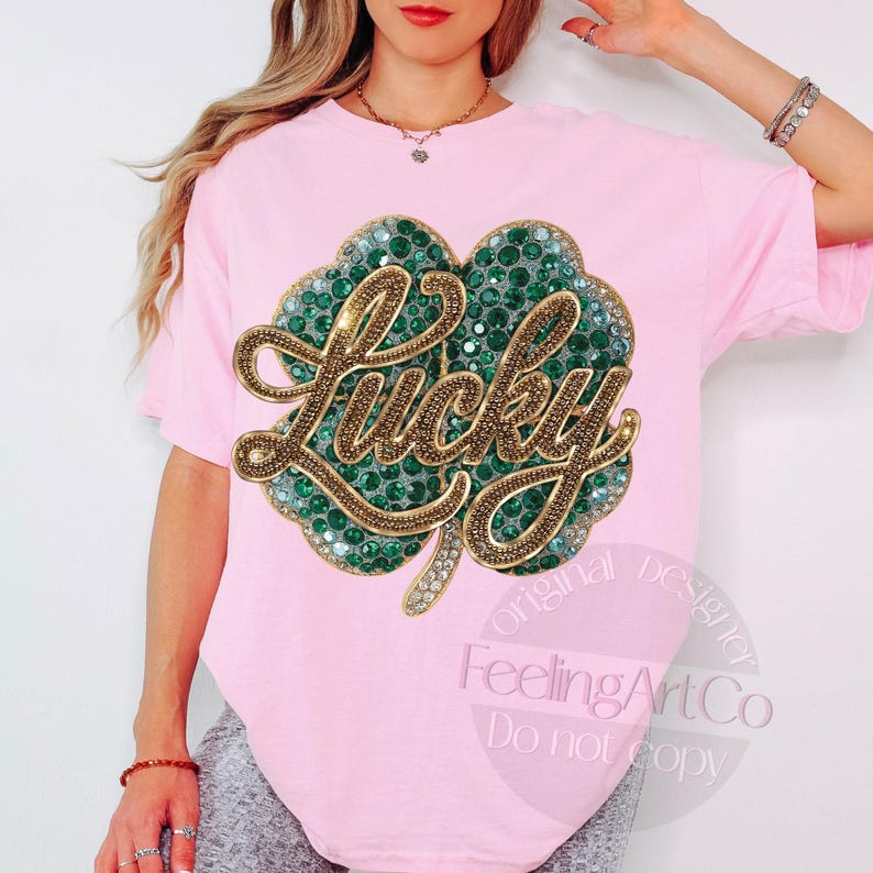 May include: A light pink t-shirt featuring a large, embellished shamrock design. The shamrock is filled with green rhinestones and outlined with gold-colored script that spells "Lucky". The shirt has short sleeves.