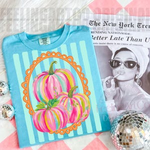 K&ouml;nnte beinhalten: Ein hellblaues T-Shirt mit vertikalen Streifen und einem dekorativen orangefarbenen Rahmen. Im Rahmen befinden sich drei bunte K&uuml;rbisse mit rosa, gelben und gr&uuml;nen Streifen. Der Zeitungstext lautet "Better Late Than U".