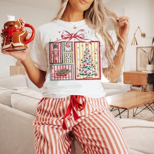 May include: White t-shirt with a Christmas-themed graphic featuring a decorated tree, Santa, and a nutcracker. The person is also wearing red and white striped pajama pants and holding a gingerbread man mug.