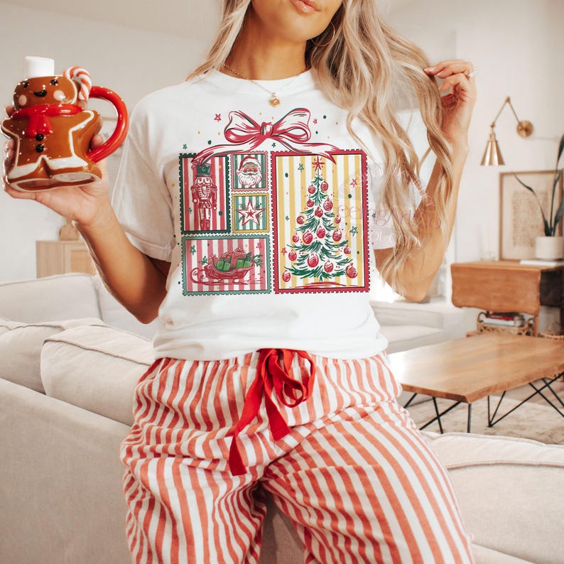 May include: White t-shirt with a Christmas-themed graphic featuring a decorated tree, Santa, and a nutcracker. The person is also wearing red and white striped pajama pants and holding a gingerbread man mug.