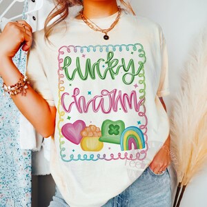 K&ouml;nnte beinhalten: Cremefarbenes T-Shirt mit einem farbenfrohen "Lucky Charm"-Aufdruck. Das Design umfasst ein Herz, einen Goldtopf, ein Kleeblatt und einen Regenbogen. Der Text ist in Gr&uuml;n und Rosa gehalten und mit einem wellenf&ouml;rmigen, farbenfrohen Rand umrandet. Ein T-Shirt zum St. Patrick's Day.