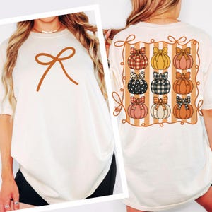 Puede incluir: Camiseta color crema con un lazo marrón en la parte delantera. La parte trasera de la camiseta muestra un diseño de nueve calabazas con lazos, cada una con un patrón y color diferente, sobre un fondo a rayas.