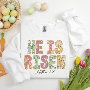 May include: White sweatshirt with a floral design that says "He is Risen" with the scripture reference "Matthew 28:6" underneath.