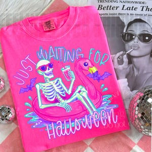 Peut inclure: T-shirt rose vif avec un motif squelette en lunettes de soleil sur un bou&eacute;e flamant rose, tenant une boisson. Le texte dit "Just Waiting for Halloween". Des chauves-souris violettes et des accents bleus compl&egrave;tent le design.
