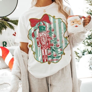May include: White t-shirt with a festive Christmas design. The graphic shows a nutcracker soldier in red and green, a Christmas tree, and a red bow. The design is framed by a green and gold border with a striped background.