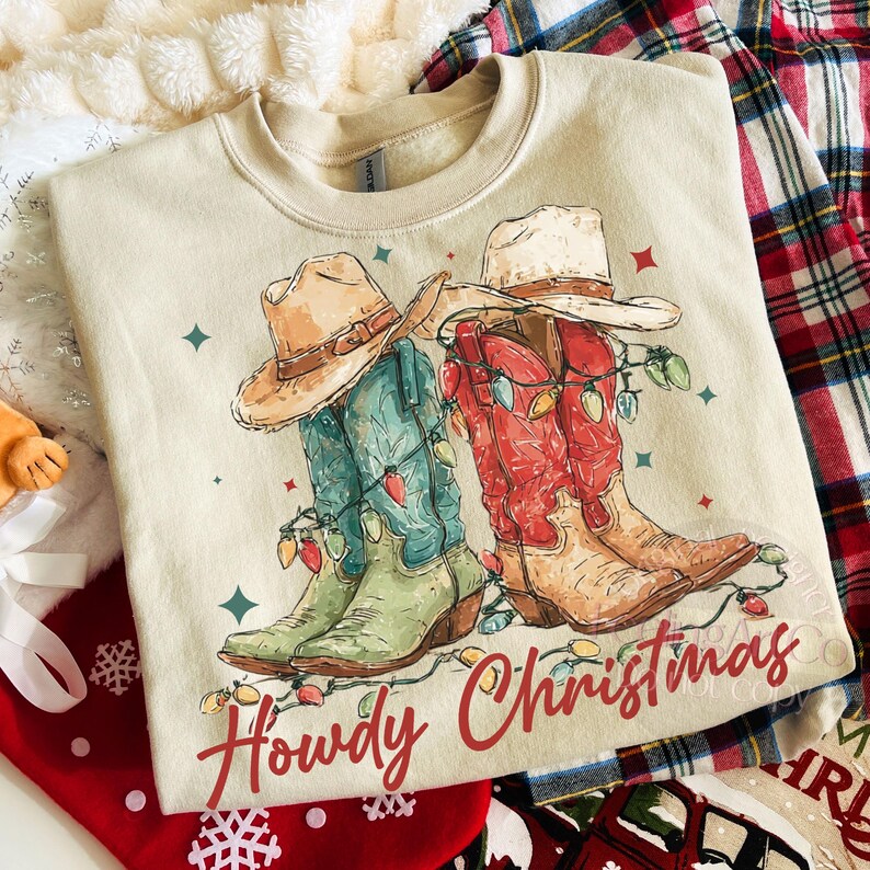May include: Beige sweatshirt with a graphic of two cowboy boots wrapped in Christmas lights and cowboy hats. The boots are green and red. The text "Howdy Christmas" is written in red script.