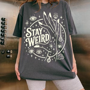May include: Dark gray t-shirt with the words "Stay Weird" in a circular design. The design includes a hand, eyes, stars, and a seashell pendant. The shirt is a casual, oversized fit.