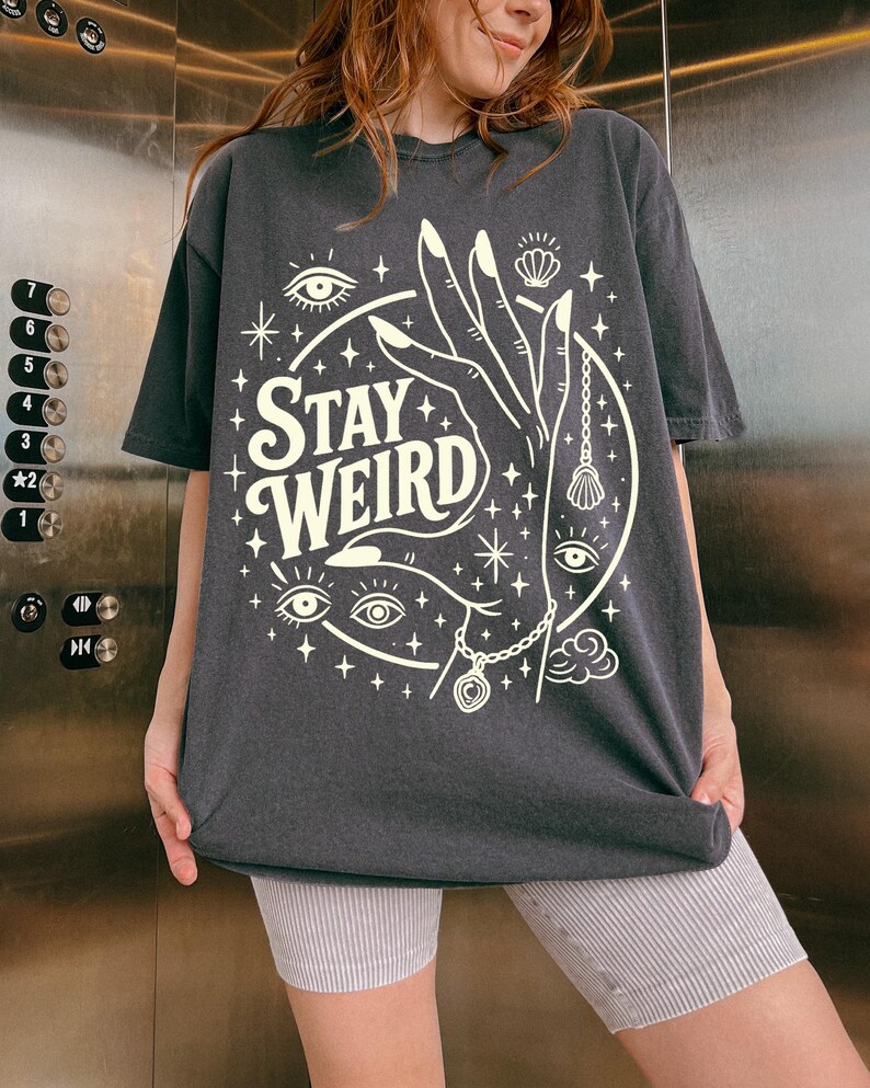 May include: Dark gray t-shirt with the words "Stay Weird" in a circular design. The design includes a hand, eyes, stars, and a seashell pendant. The shirt is a casual, oversized fit.