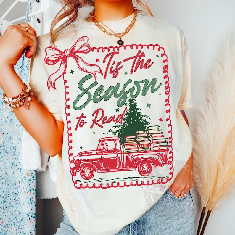 May include: Cream t-shirt with a red truck graphic carrying books and a Christmas tree. The text reads "Tis The Season to Read" in green and red. A red bow and decorative border complete the design.