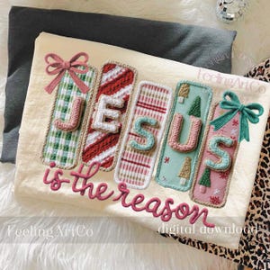 May include: A cream-colored textile features the words "JESUS is the reason" in a decorative script. The word "JESUS" is spelled out vertically with colorful, patterned panels and bows. The design includes Christmas tree and snowflake motifs.