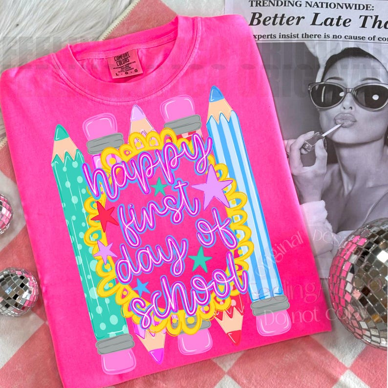 Puede incluir: Una camiseta de color rosa brillante con un estampado gr&aacute;fico de l&aacute;pices coloridos y un borde festoneado amarillo que rodea el texto "happy first day of school" en letras cursivas moradas, acentuado con peque&ntilde;as estrellas rosas, azules y moradas.