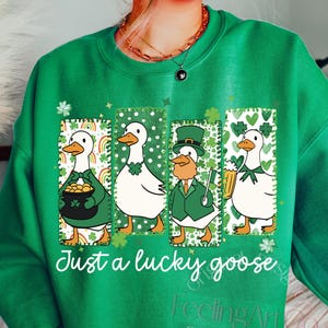 May include: Green sweatshirt with a St. Patrick's Day theme. Features four cartoon geese in rectangular frames, each with a different design, including rainbows, shamrocks, and a leprechaun. The text "Just a lucky goose" is printed below the images.