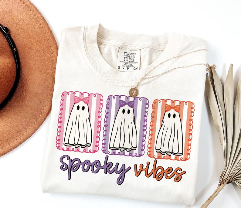 May include: Cream-colored t-shirt with a Halloween-themed graphic. The design features three cartoon ghosts with bows, set in colorful frames, and the words "Spooky Vibes" in purple and orange.